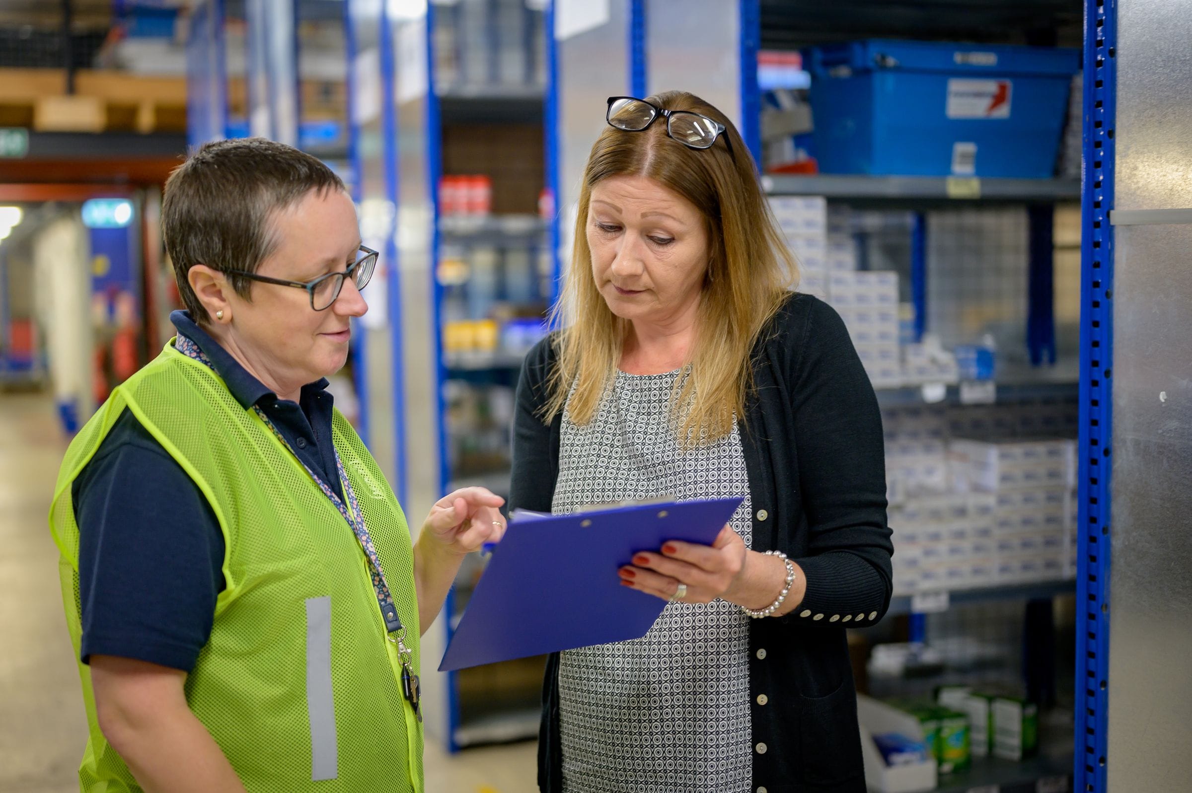 A buyer and a warehouse worker checking stock against a list between racks