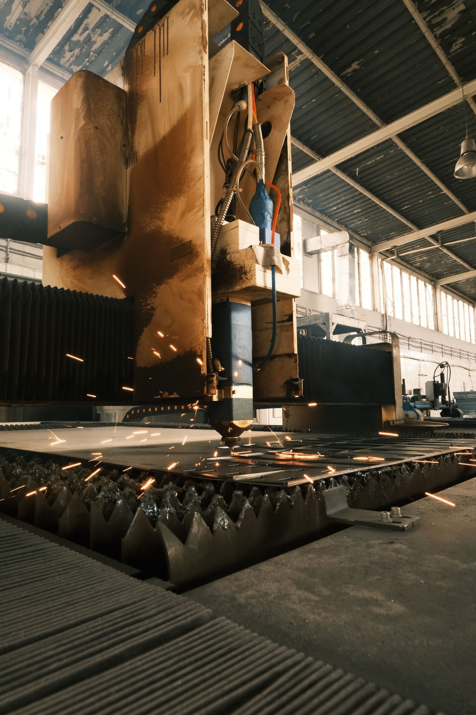 A laser cutting head over a steel plate in a tall, sunlit production hall